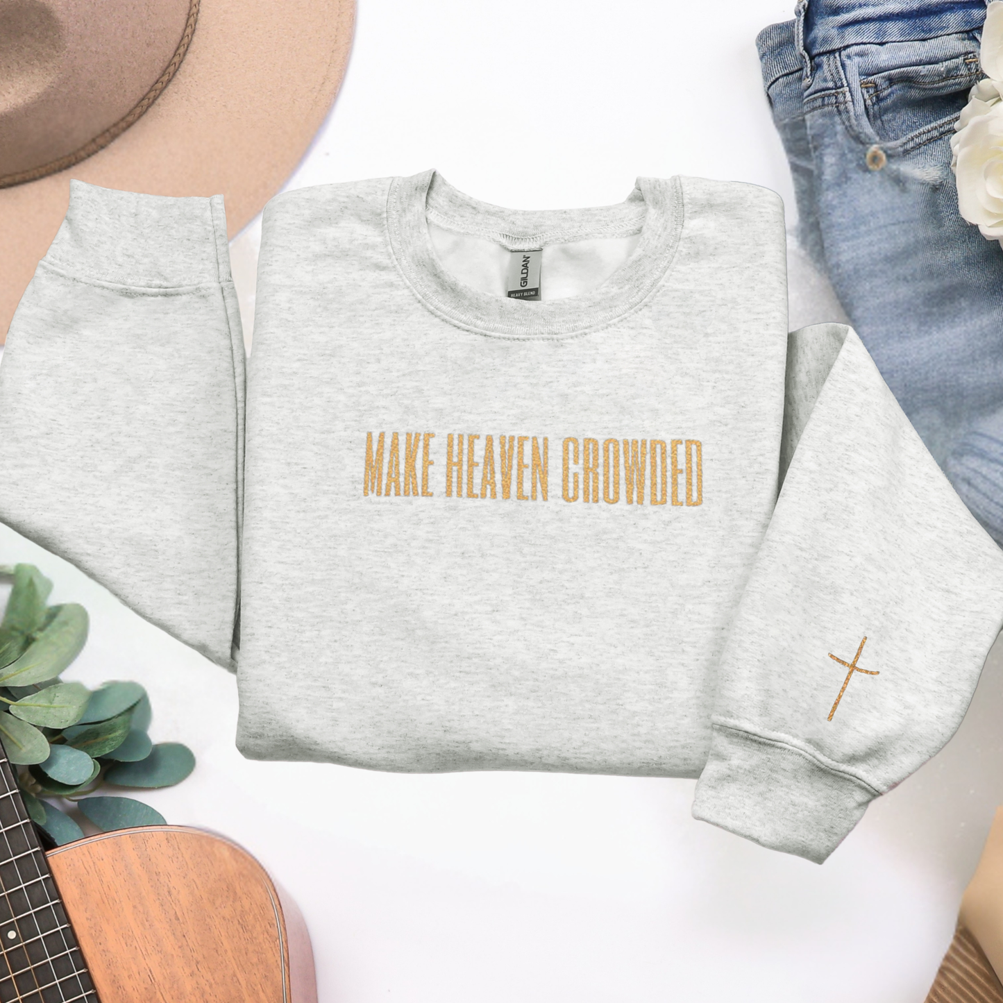 Gray sweatshirt with 'MAKE HEAVEN CROWDED' text and cross design on a white surface with accessories.