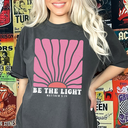 Person wearing a black t-shirt with a pink sun graphic and 'Be the Light' text, standing in front of a colorful album cover wall.