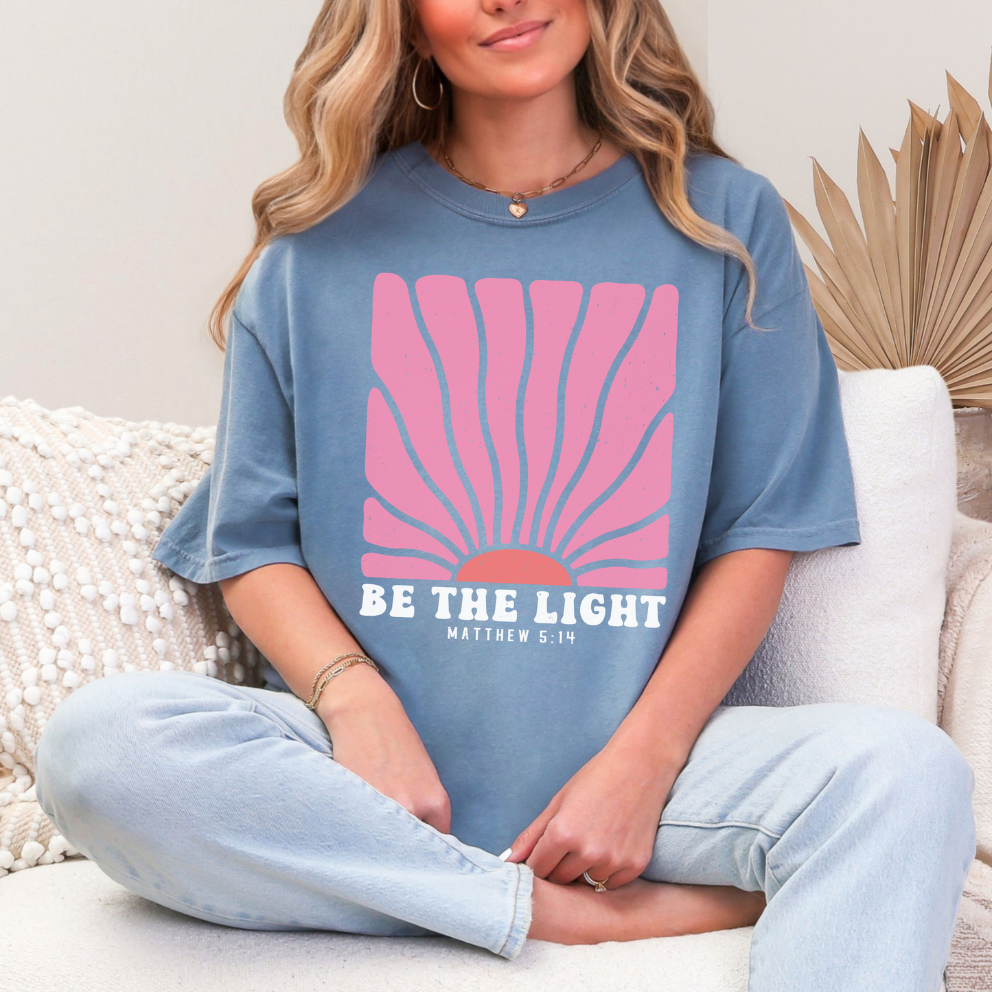 Person wearing a blue t-shirt with a pink graphic and 'Be the Light' text, sitting on a couch.