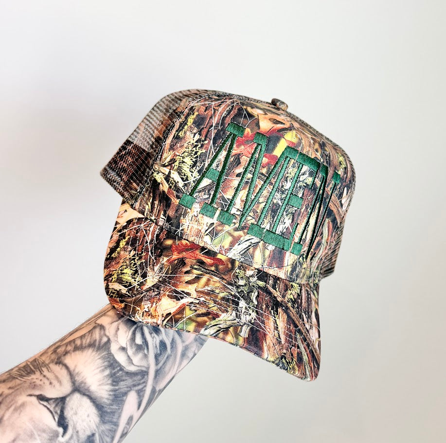 Camouflage-patterned cap with embroidered text 'AMEN.' on a light gray background