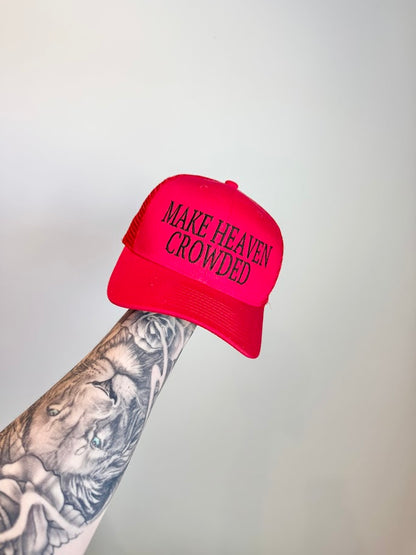 Red hat with 'MAKE HEAVEN CROWDED' text held by a tattooed arm against a gray background