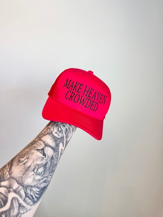 Red hat with 'MAKE HEAVEN CROWDED' text held by a tattooed arm against a gray background